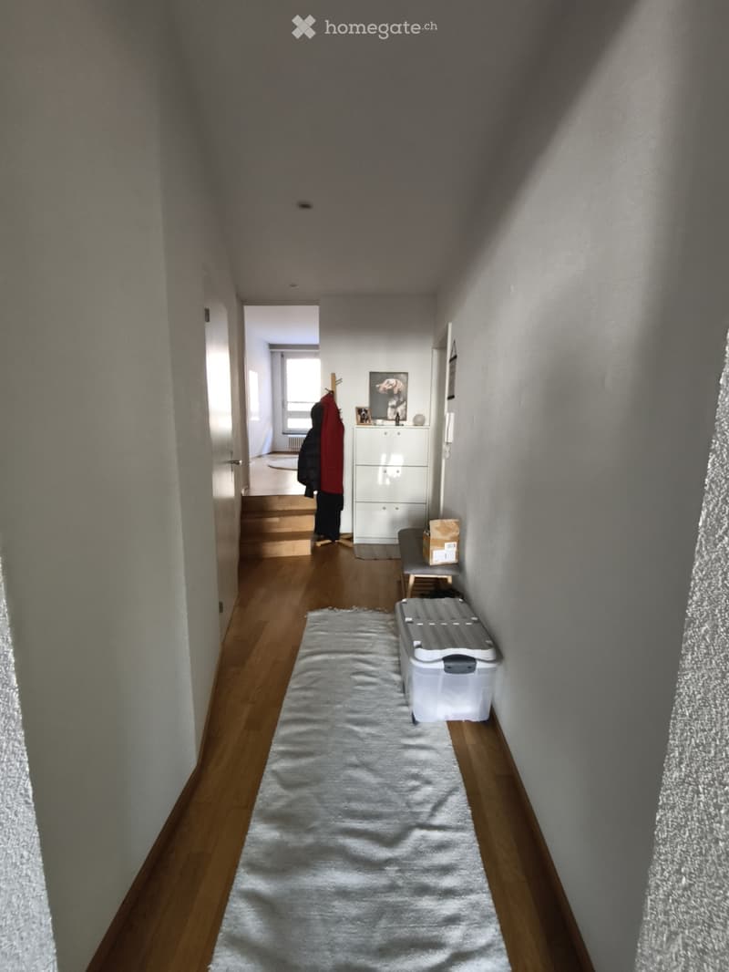 A narrow hallway with wooden flooring, white walls, and a white rug in the center. A small bench and storage unit are visible at the end of the hallway.
