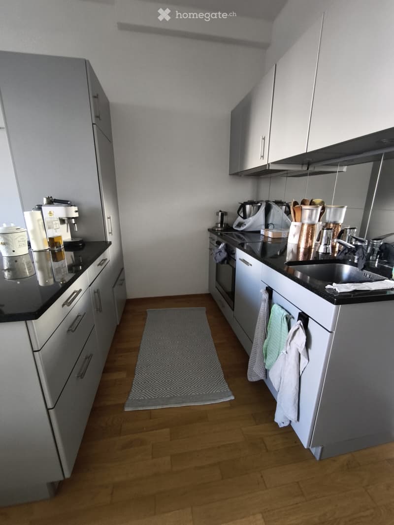 Modern kitchen with grey cabinets, black countertops, wooden flooring, and various appliances including a coffee machine, blender, and oven.