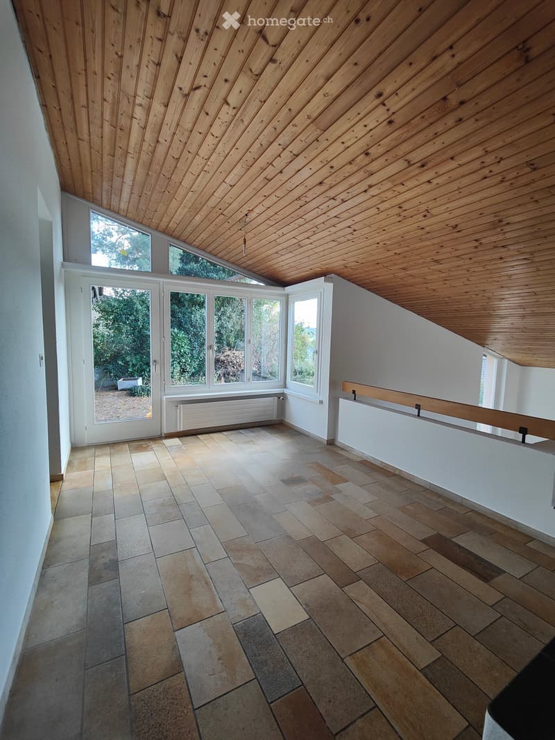 Bright, empty room with wooden slanted ceiling, large windows, tiled floor, and white walls.