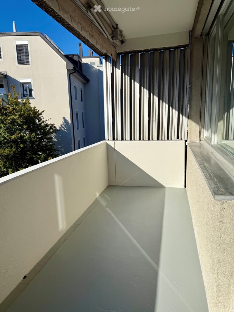 Balcony with white walls, a retractable awning, and a view of neighboring buildings and greenery.
