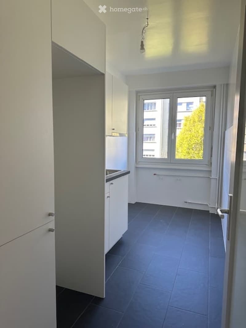 Empty kitchen with white cabinets, dark tiled floor, and a window overlooking a building and tree.