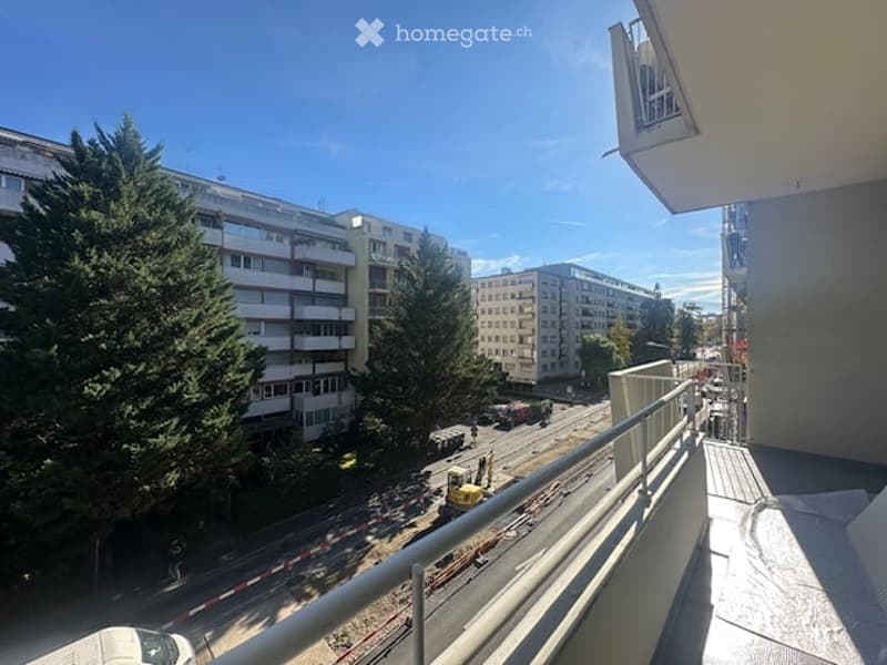 Balcony overlooking a city street with buildings, trees, and a clear blue sky.