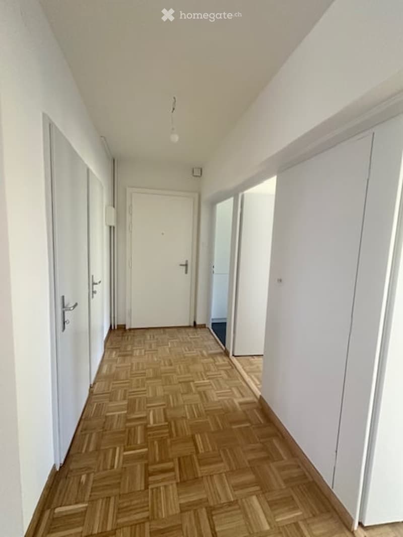 A clean, well-lit hallway with wooden herringbone flooring, white walls, and several closed doors.