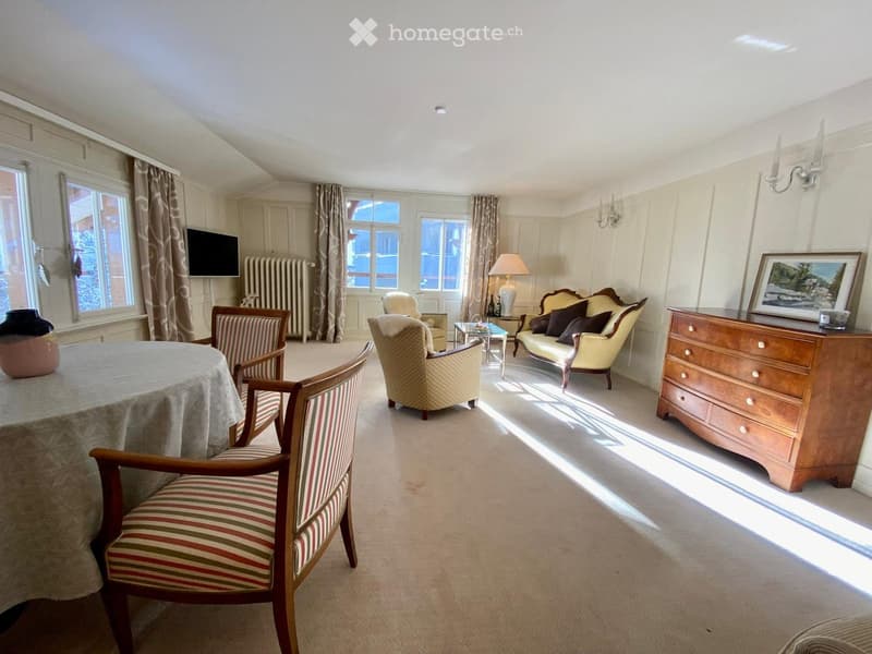 Bright living room with large windows, cream carpet, and classic furniture including a sofa, armchairs, a coffee table, and a dining table with chairs.