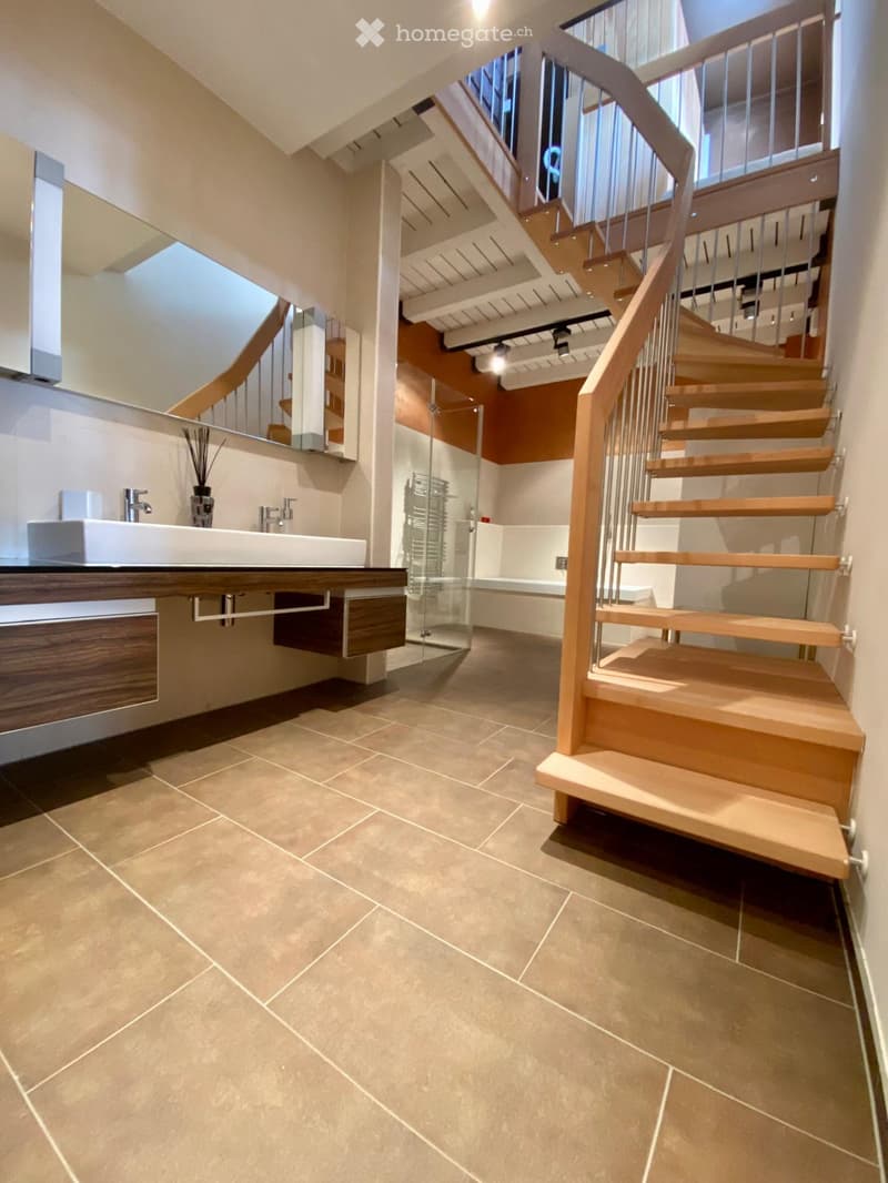 Modern hallway with a wooden staircase, beige tiled floor, and a small bathroom area featuring a sink, mirror, and glass shower enclosure.