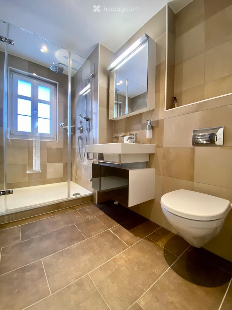 Modern bathroom with beige tiles, a white sink, toilet, mirror, and a glass-enclosed shower with a window.
