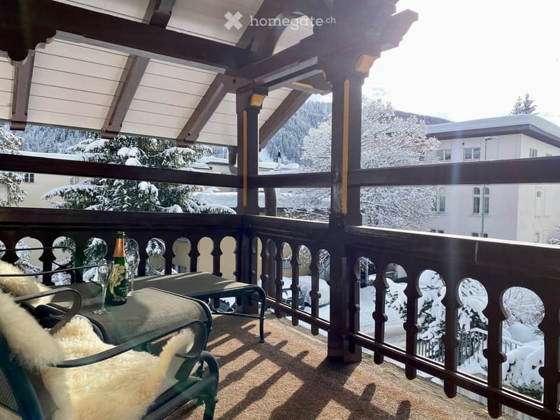A wooden balcony with a view of snow-covered trees and buildings, featuring a chair with a fur throw, a small table with a bottle and wine glass.