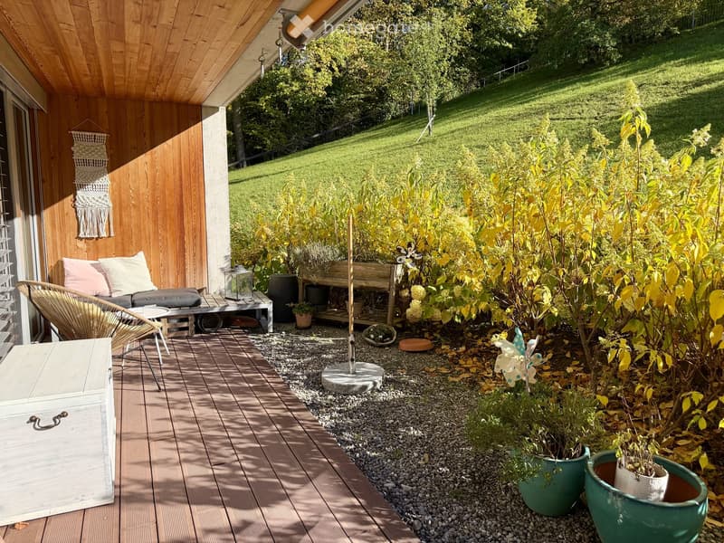 A wooden deck area with a bench, cushions, a small table, and potted plants, surrounded by a gravel garden with shrubs and trees, overlooking a grassy hill.