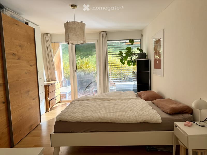 Modern bedroom with a double bed, nightstand, lamp, wardrobe, and large windows with curtains. A plant is placed near the window, and a picture frame hangs on the wall.