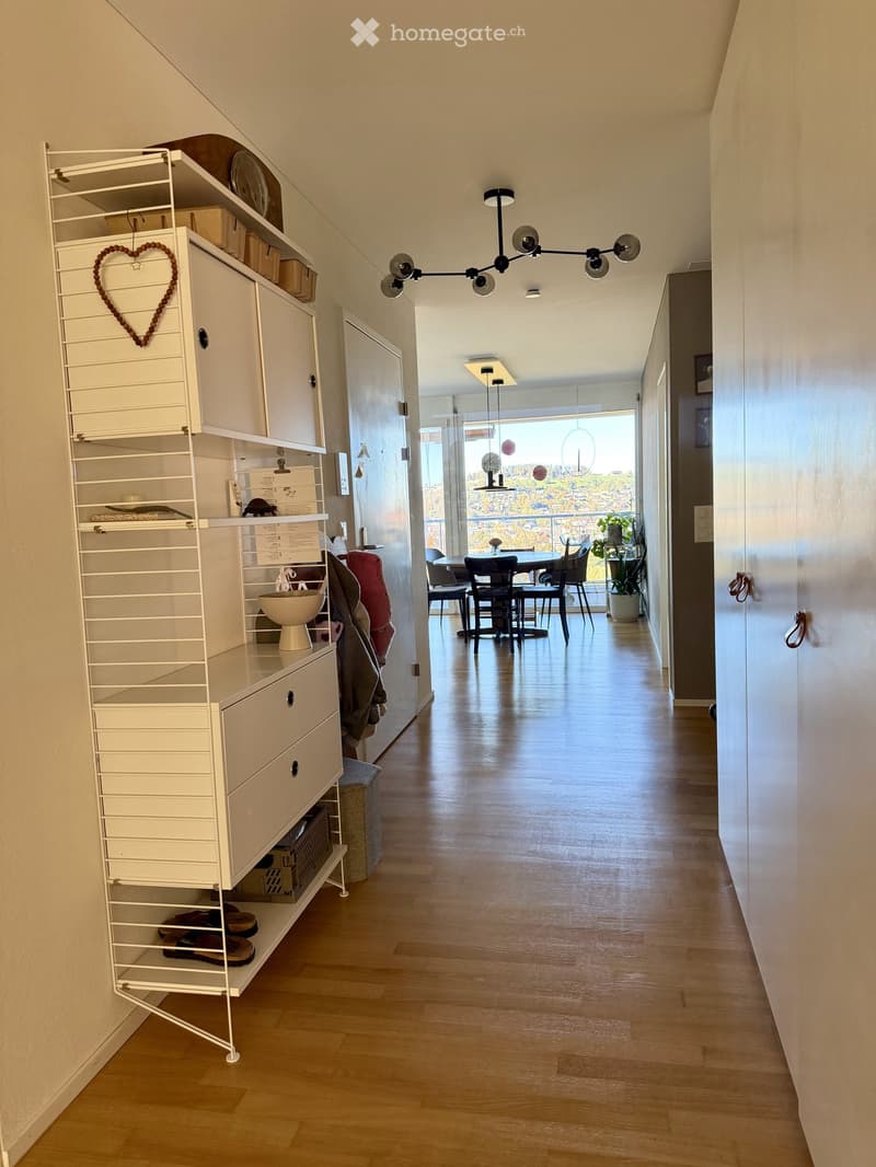 A modern hallway with wooden flooring, white shelving unit on the left, and a dining area visible at the end with a round table and chairs. Natural light streams in from a large window at the far end.