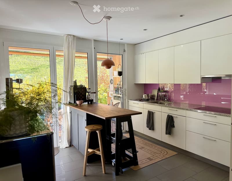Modern kitchen with white cabinets, a wooden island, and a purple backsplash. Features include a coffee machine, sink, and large windows with curtains.
