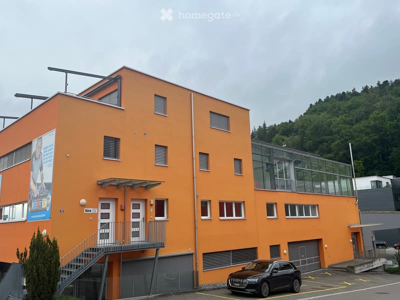 Modern two-story building with orange facade, multiple windows, and a balcony. Features a staircase leading to an upper level and a parked car in front.