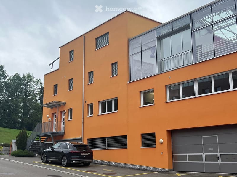 Modern orange-colored building with multiple windows, a balcony, and a garage. A car is parked in front of the building.