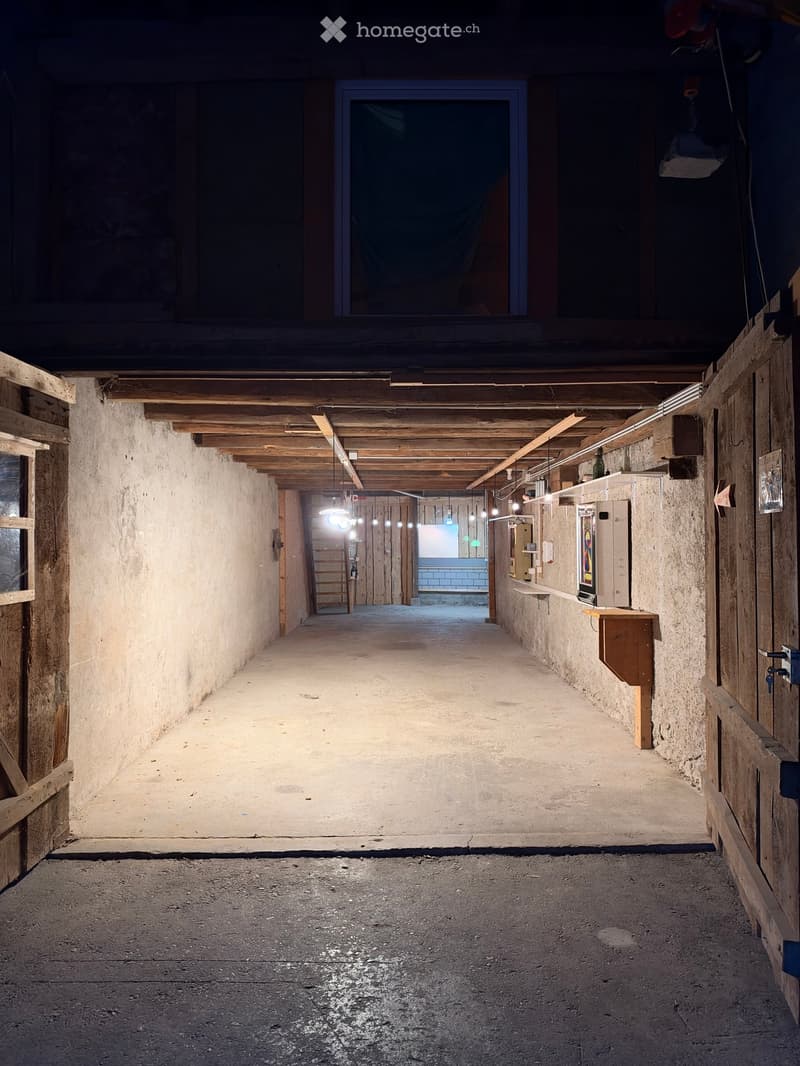 A long, narrow corridor with exposed wooden beams and concrete walls, featuring a series of doors and a small bench on the right side.