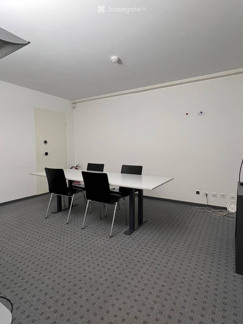 A small, modern meeting room with a white table, four black chairs, and a fire extinguisher on the table. The room has white walls, a grey carpet, and a white door.