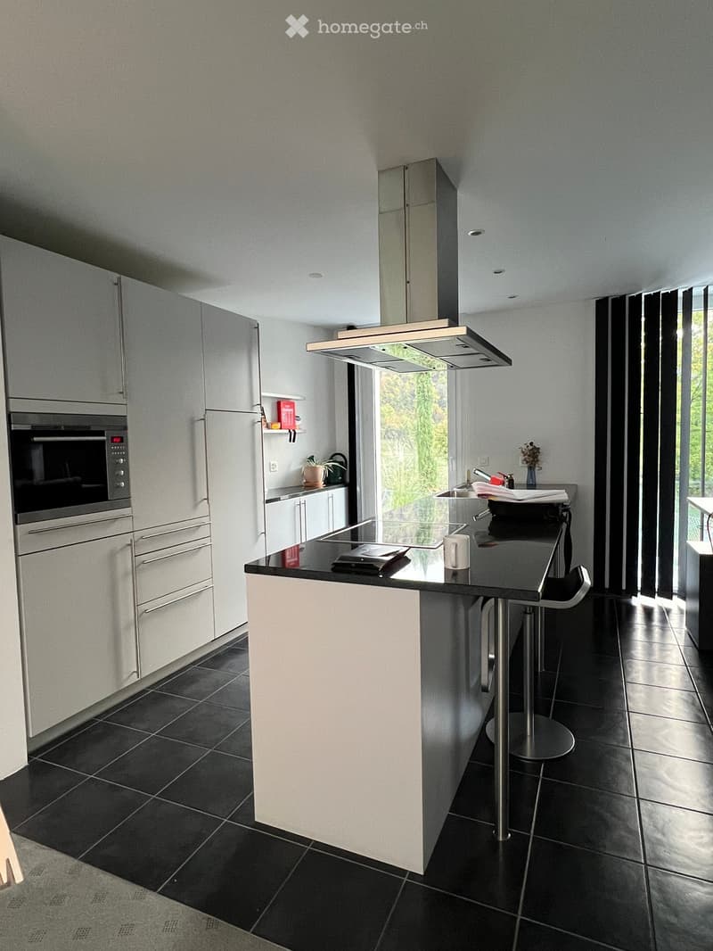 Modern kitchen with white cabinetry, black countertops, and stainless steel appliances. Features an island with a sink, a stool, and a range hood above the stove.