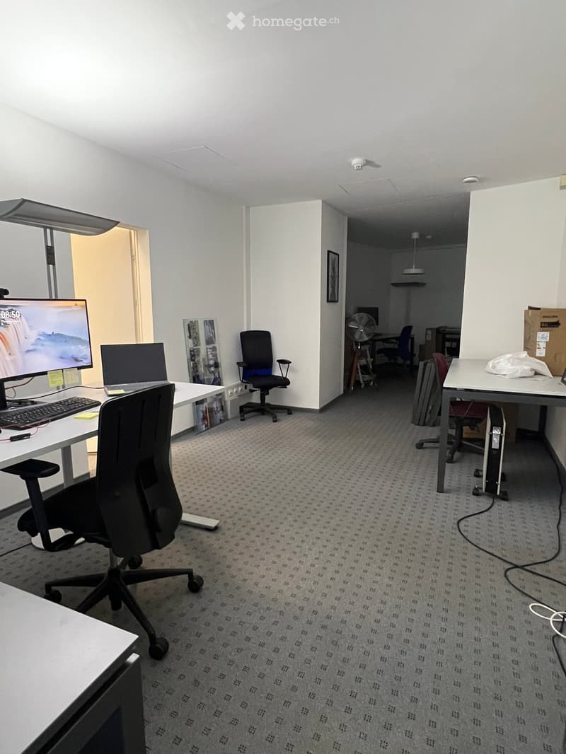 Office space with desks, chairs, a laptop, a screen, a keyboard, and a fan. The room has white walls and a carpeted floor.