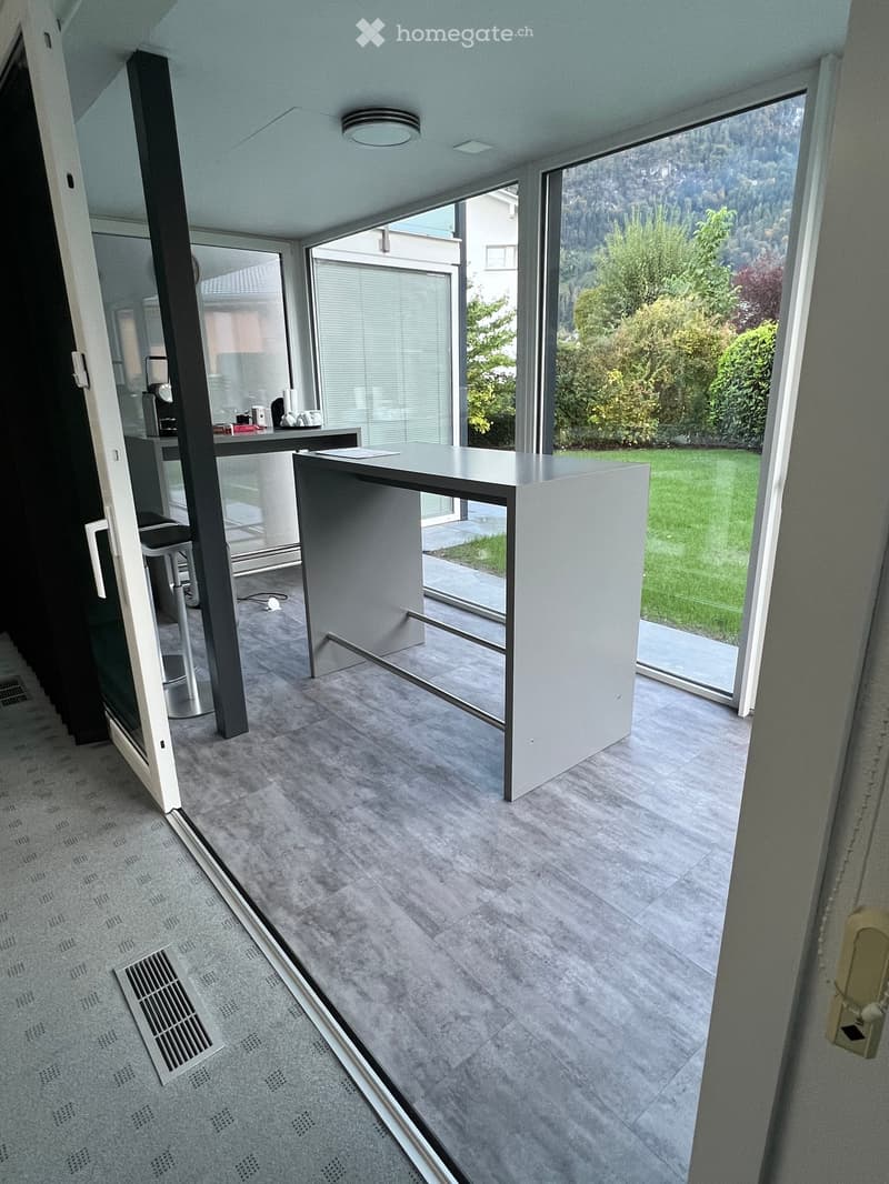 Modern, open-plan room with grey carpet, large glass doors leading to a garden, a small table, a stool, and a coffee machine visible in the background.