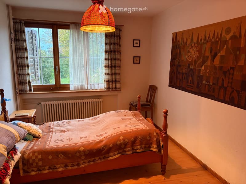 Cozy bedroom with wooden bed, patterned bedspread, wooden chair, small table, radiator, large window with curtains, and artwork on the wall.