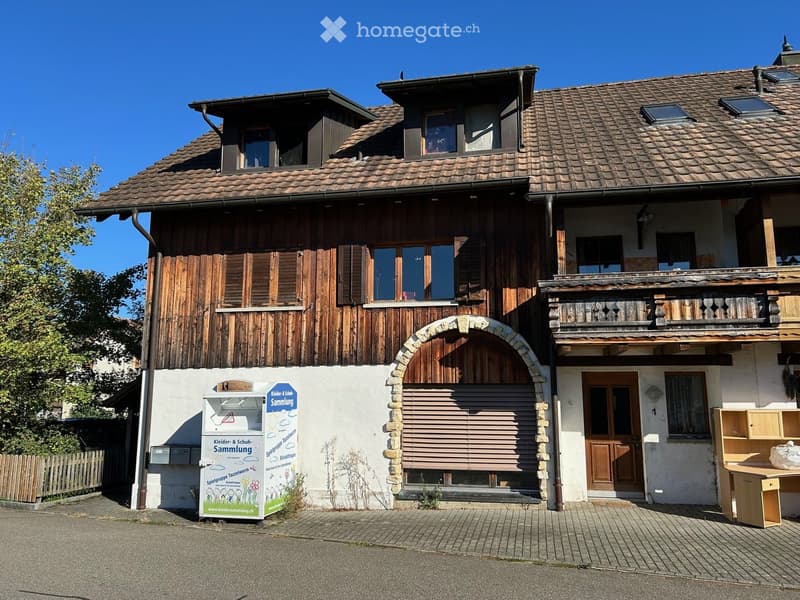 Two-story house with wooden facade, tiled roof, and balcony on the upper level. Features include a stone archway entrance, wooden shutters, and a small signboard on the left side.