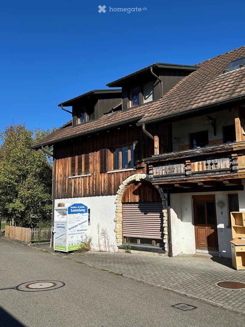 Two-story house with a tiled roof, wooden shutters, and a stone archway entrance. The building features a balcony on the upper floor and a signboard in front.
