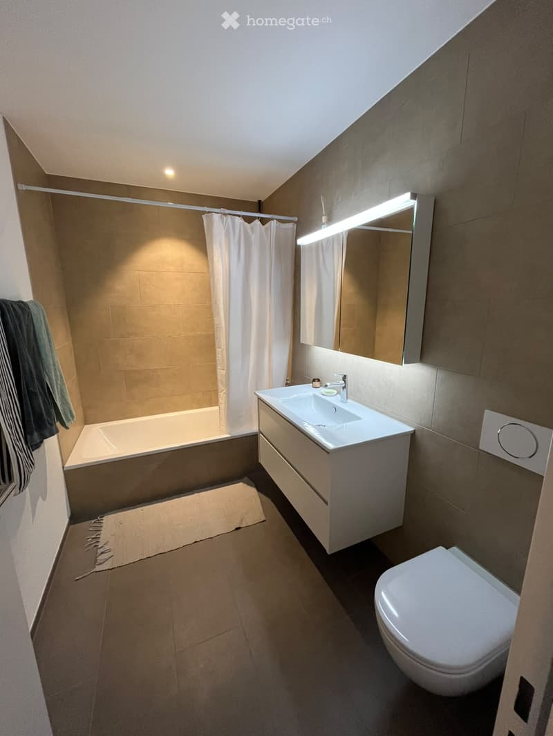 Modern bathroom with a bathtub, sink, toilet, and mirror. Features a built-in vanity with a sleek design, a shower curtain, and a towel on the floor.