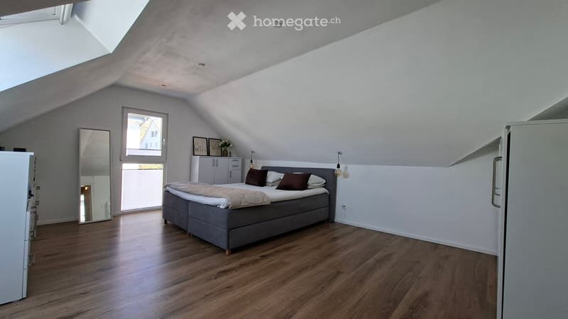 Modern attic bedroom with wooden flooring, large window, grey bed with brown pillows, mirror, small cabinet, and decorative items on top.