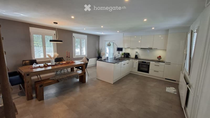Modern kitchen with white cabinets, a central island, and a dining area featuring a wooden table and bench. The space includes an oven, sink, and various appliances.