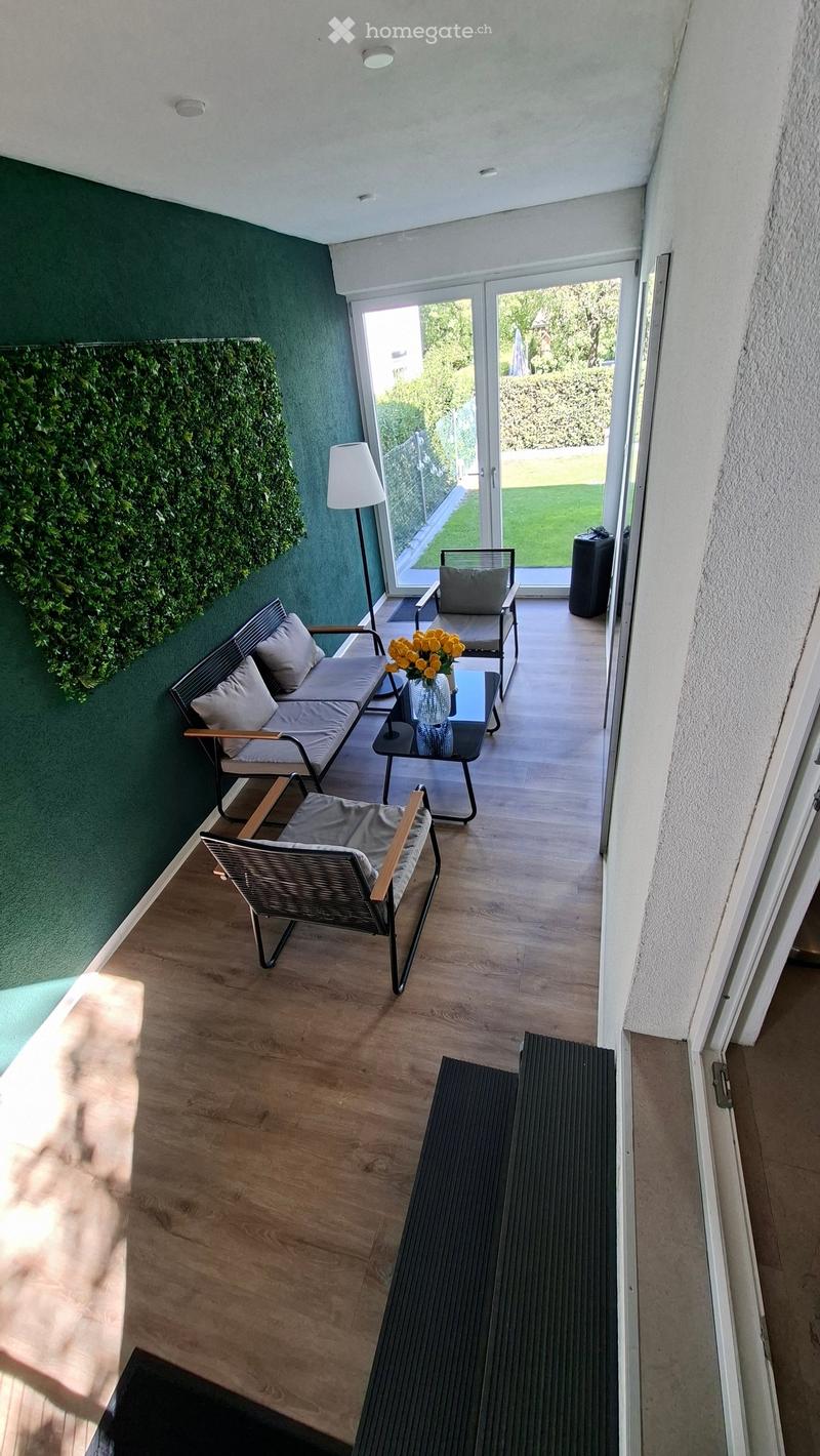 Balcony with wooden flooring, green walls, and a seating area including a sofa, chairs, a small table with a vase of yellow flowers, and a floor lamp.