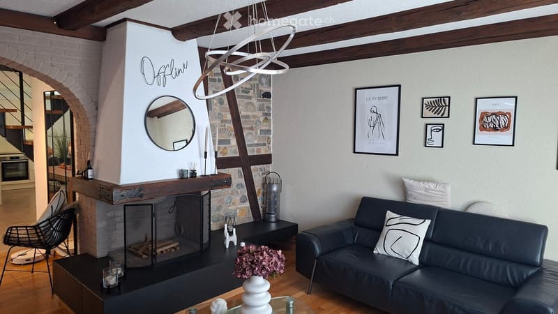 Modern living room with exposed wooden beams, a stone fireplace, a black leather sofa, a wicker chair, a coffee table, and several decorative items including candles, a vase with flowers, and artwork on the walls.