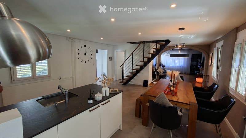 Modern kitchen with a black countertop, sink, and faucet. A wooden dining table with black chairs is visible, along with a staircase leading to another level.