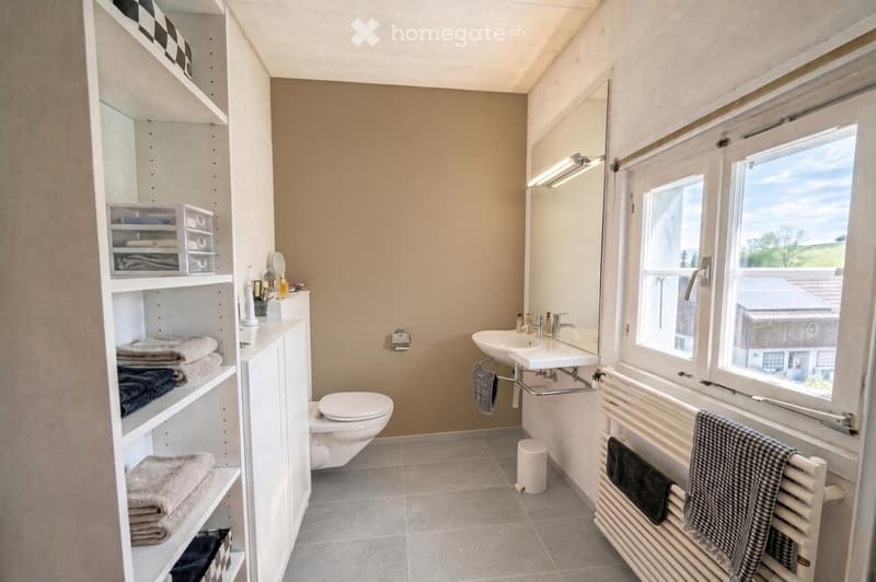 Modern bathroom with a toilet, sink, mirror, radiator, and shelves holding towels and toiletries. The room features large windows with a view of the outdoors.