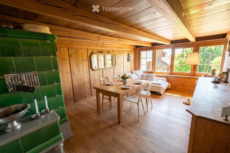 Cozy wooden room with exposed beams, green tiled stove, dining table with chairs, sofa, and large windows letting in natural light.