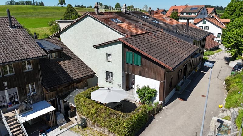 Aerial view of a residential area with multiple houses, featuring red-tiled roofs, green shutters, and a mix of modern and traditional architecture. The houses are surrounded by greenery and a scenic countryside backdrop.