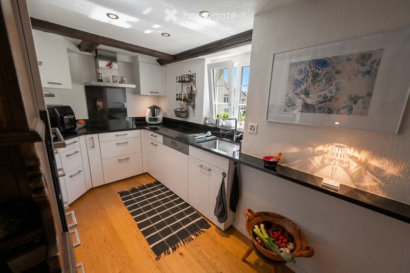 Modern kitchen with white cabinets, black countertops, stainless steel appliances, and a wooden floor. Features include a sink, faucet, blender, framed picture, table lamp, and a bowl of vegetables on the counter.