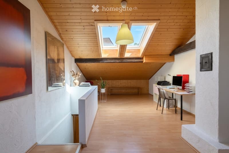 Bright attic room with wooden floors and slanted wooden ceiling, featuring a desk with a computer, a hanging lamp, a small bench, and decorative items on a shelf.