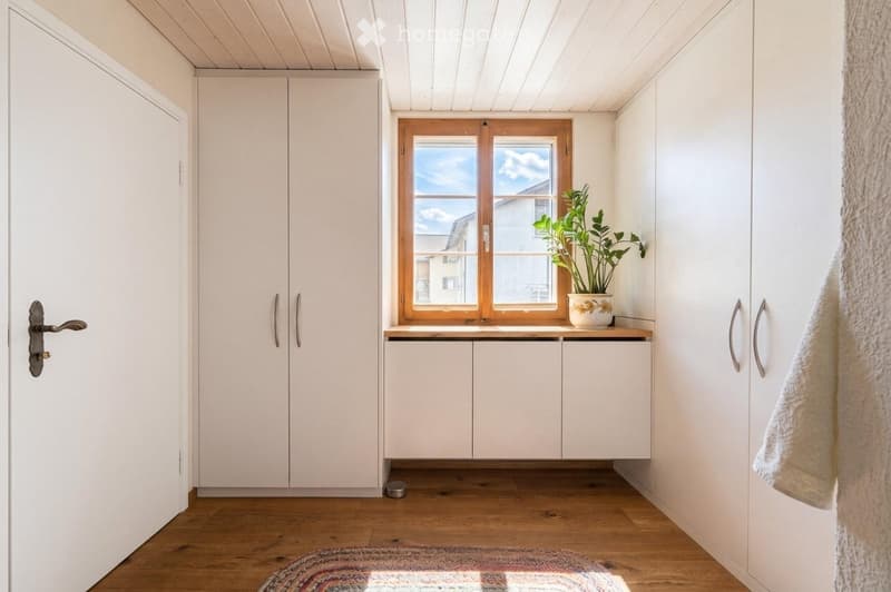 A small, well-lit room with white cabinets, a wooden floor, a window with a plant on the sill, and a towel hanging on the wall.