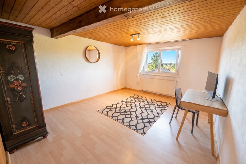 Bright room with wooden flooring, a large window with white curtains, a patterned rug, a wooden desk with a chair, a screen, and a decorative wardrobe.
