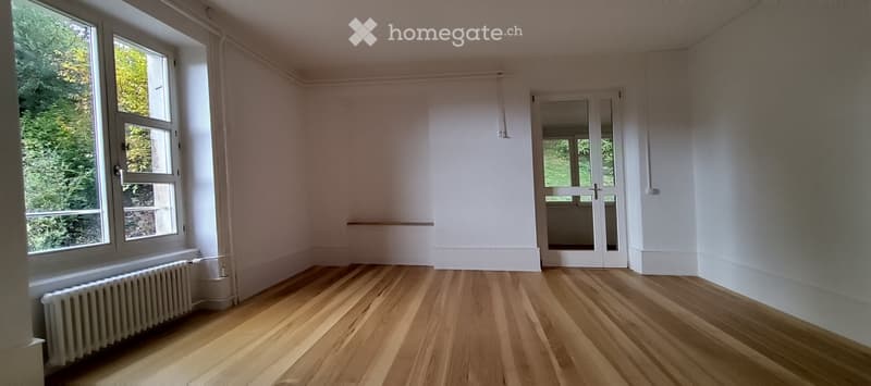 Empty room with light wooden flooring, white walls, large windows, and a radiator. No furniture or equipment present.