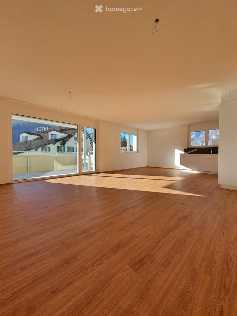 Empty room with wooden flooring, large windows, and a small kitchen area with cabinets visible in the background.