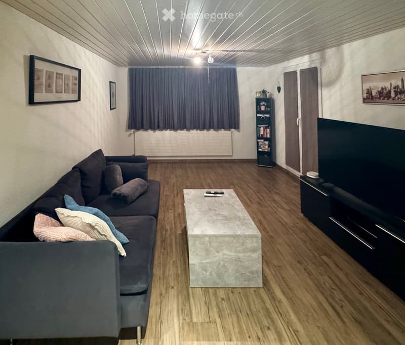 Living room with a dark grey sectional sofa, a marble coffee table, a TV stand with a flat-screen TV, a small bookshelf, and framed artwork on the walls.