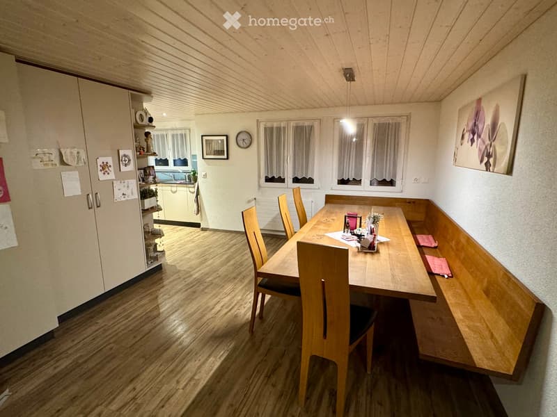 Dining area with a wooden dining table, chairs, a built-in bench, and a small kitchen area visible in the background.