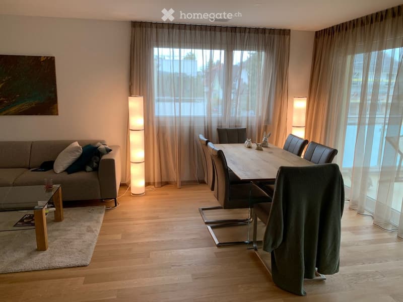 Modern living and dining area with a grey sofa, coffee table, dining table with chairs, and floor lamps. The space features large windows with sheer curtains and a light wooden floor.