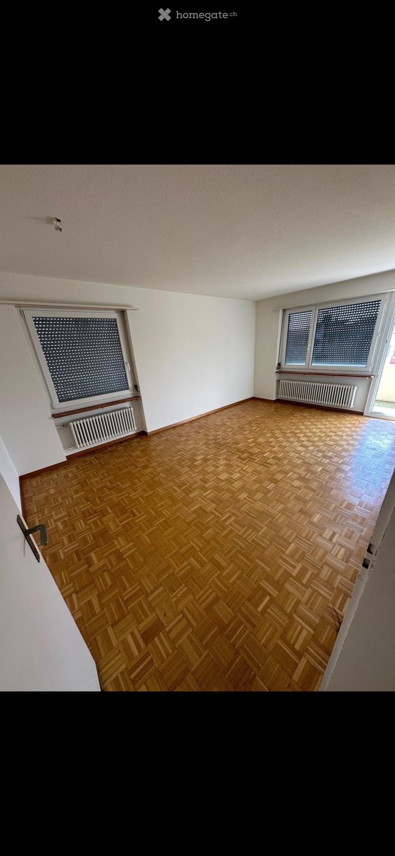 Empty room with wooden herringbone flooring, white walls, and large windows with blinds. Radiators are visible beneath the windows.