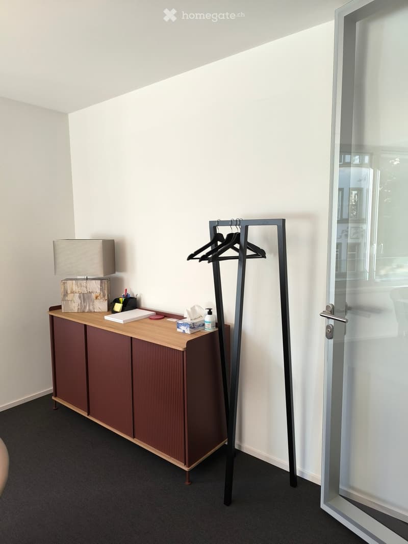 A small, modern room with a wooden table, a lamp, a coat rack, and a glass door leading outside.