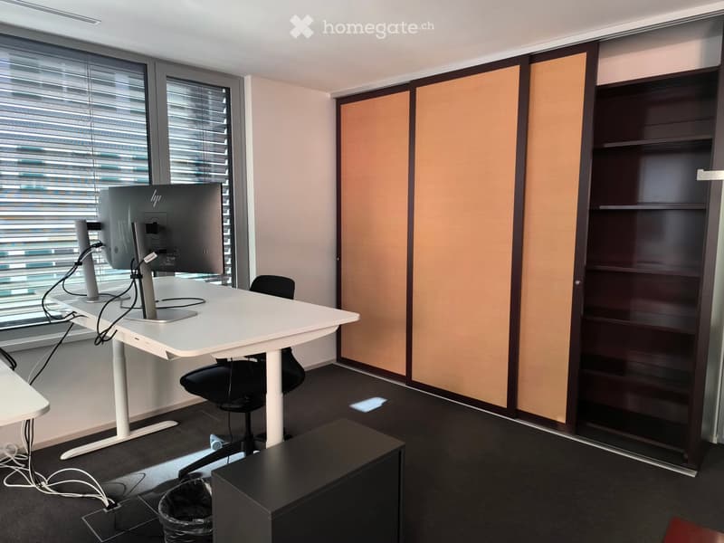 Modern office space with a white standing desk, ergonomic chair, dual monitors, and a dark wooden shelving unit.