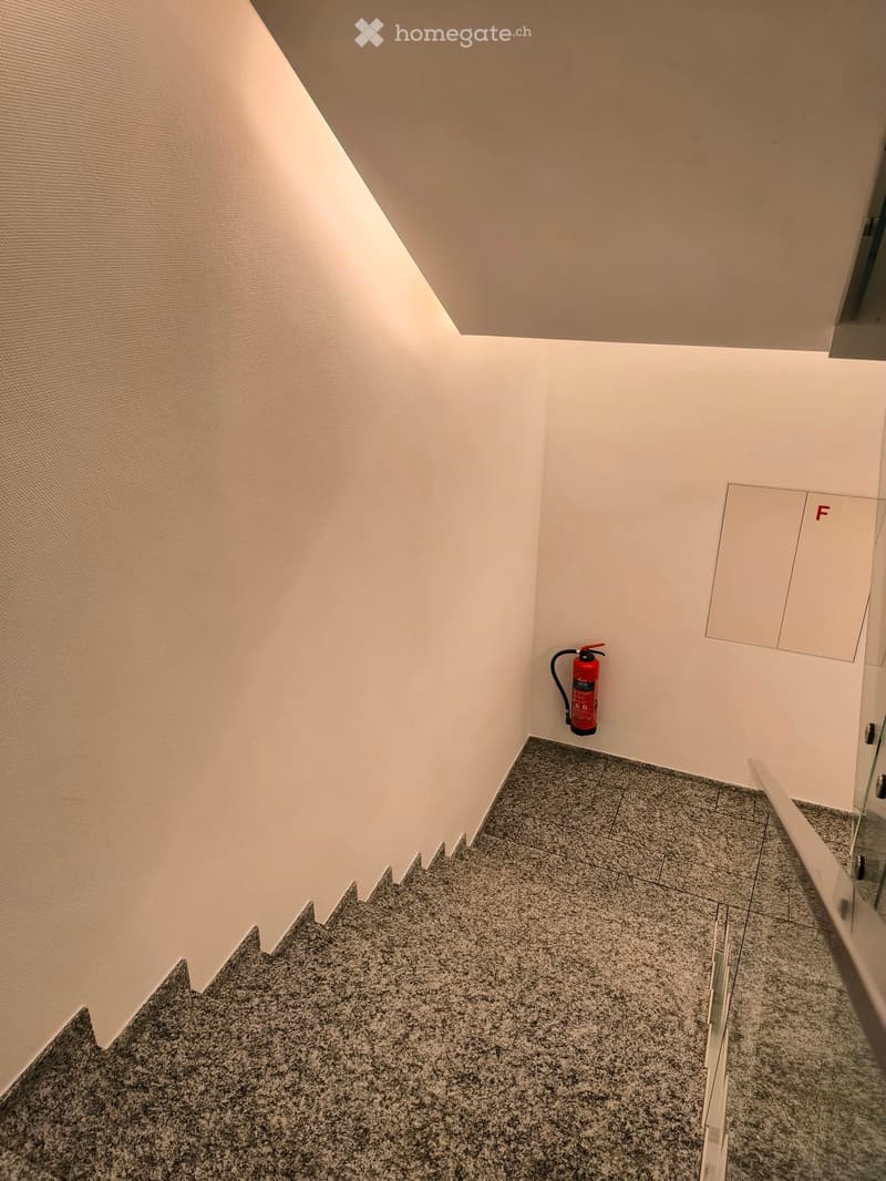 A modern hallway with white walls, grey tiled floor, and a red fire extinguisher mounted on the wall.