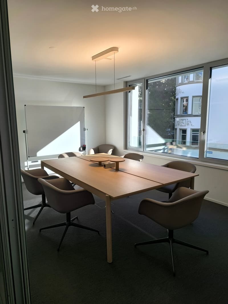 Modern office meeting room with a long wooden table, several chairs, a large screen, and large windows with a view of the outside.