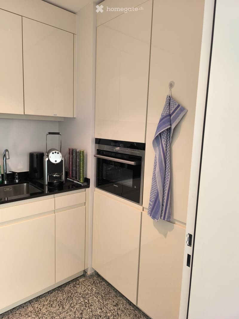 Modern kitchen with light-colored cabinets, stainless steel oven, coffee machine, and a cup rack on the counter.