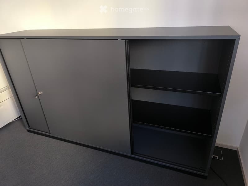 Empty room with a grey cabinet featuring a closed door and open shelves, placed against a white wall with a grey carpeted floor.